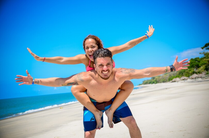 Une femme sur le dos d’un homme qui ont tout les deux les bras en l’air sur une plage