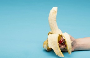 Une main qui tient une banane épluchée à la base avec un coulis blanc qui sort du haut de la banane pour simuler une éjaculation sans masturbation.