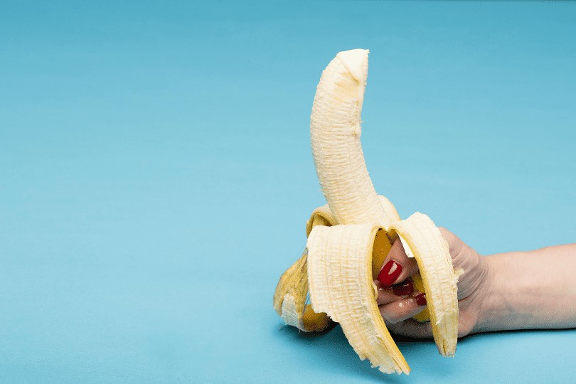 Une main qui tient une banane épluchée à la base avec un coulis blanc qui sort du haut de la banane pour simuler une éjaculation sans masturbation.